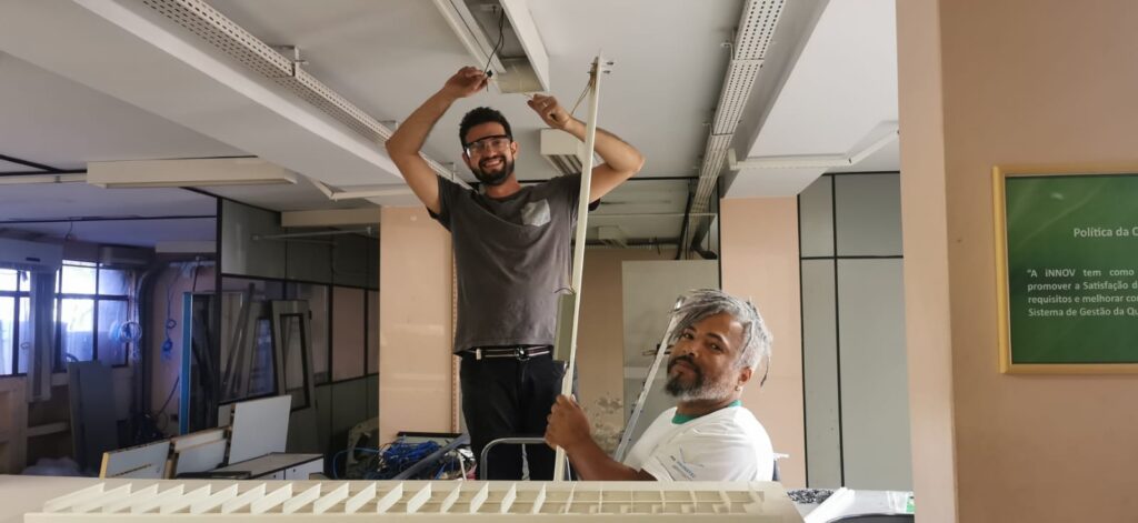 Gustavo, de óculos de proteção, instalando uma lâmpada no teto, enquanto Darlon segura a luminária