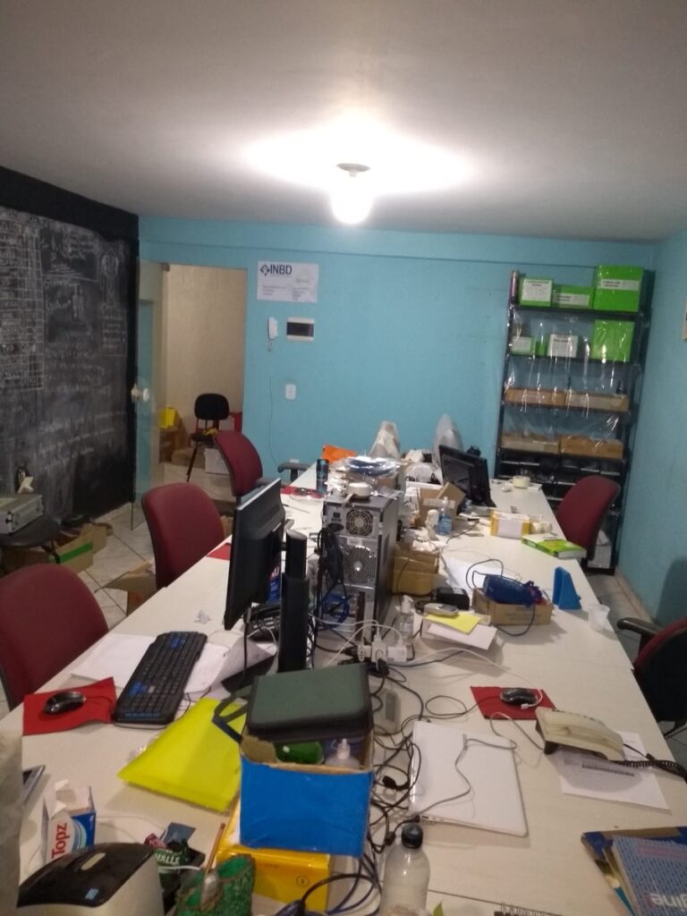 Sala pequena com uma mesa comprida cheia de equipamentos e computadores, e o quadro negro na parede ao fundo